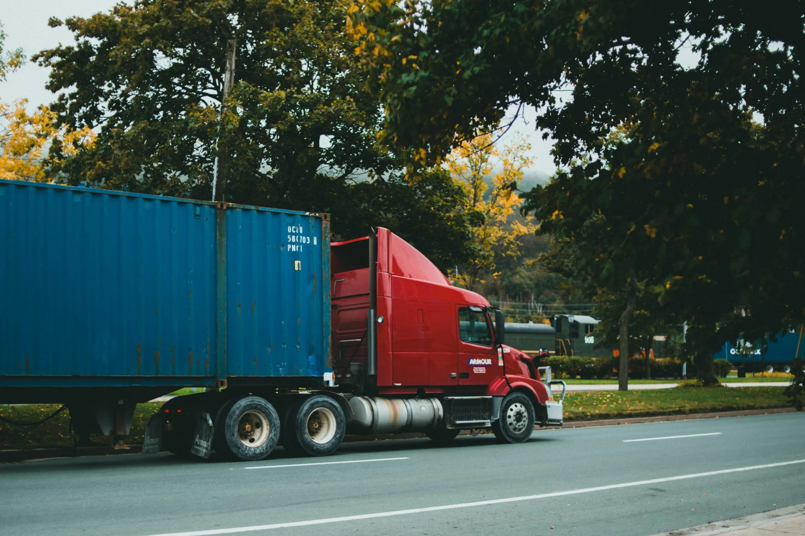 A red truck transports a blue cargo container on a scenic road,